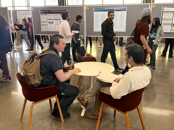 [DOM faculty (seated) compare notes on evaluating residents’ research as participants pass by looking at posters.]