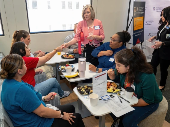 [Cynthia Campas (standing center), DOM associate administrator, and Dominica Padilla (standing right), DOM operations support manager, pass out raffle tickets for prizes to be won later to attendees of the 2025 DOM Administrative Professionals Day breakfast.]