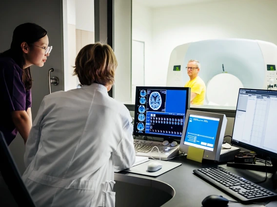[Image of two researchers looking at MRI image of brain while man in yellow shirt, visible through window in front of them, sits on MRI machine in next room.]