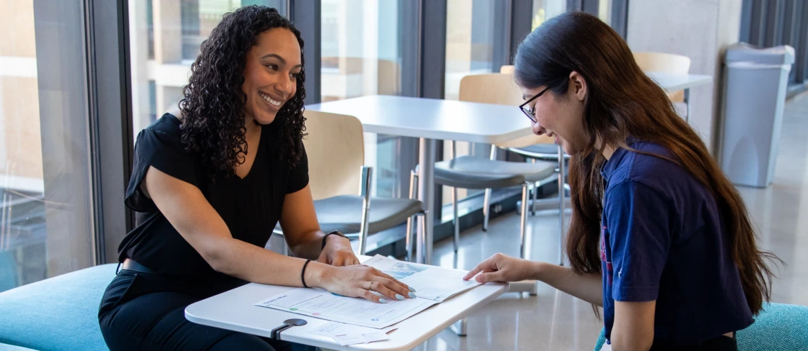 Two women seated at a table, examining a brochure together, engaged in discussion and sharing ideas.