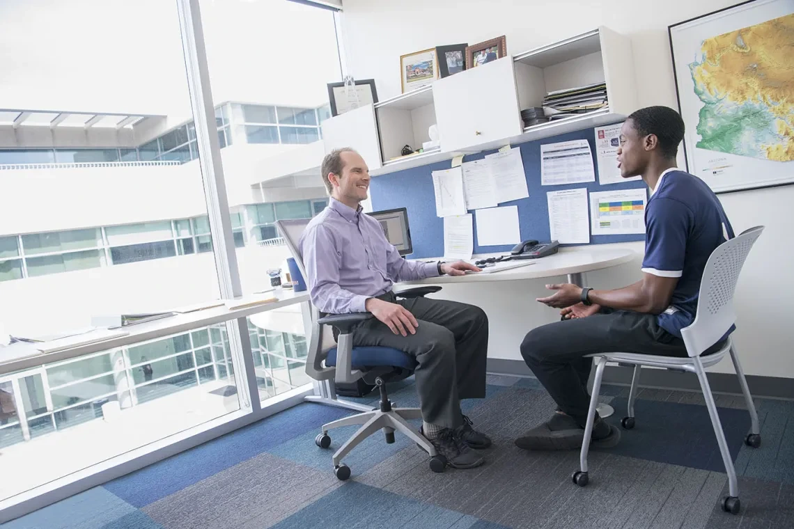Two men at an office desk, collaborating on a project with papers and a computer on th desk.