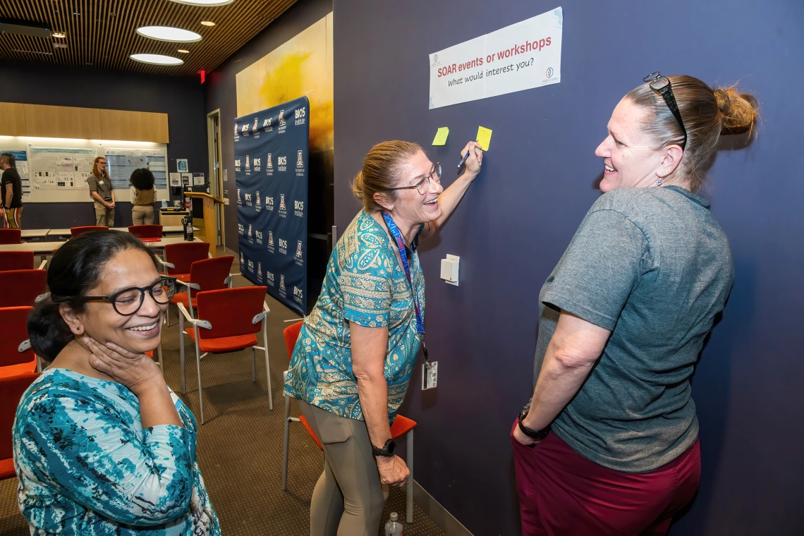 [Cardiology’s Shibani Mukerjee, PhD (left), laughs with colleagues as they come up with thoughts to add to post-its for the SOAR in DOM research forum’s “Idea Wall.”]