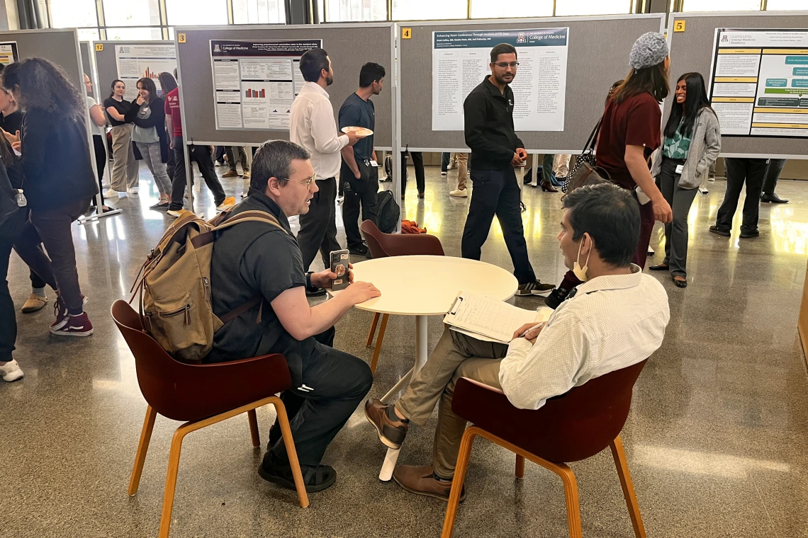 [DOM faculty (seated) compare notes on evaluating residents’ research as participants pass by looking at posters.]