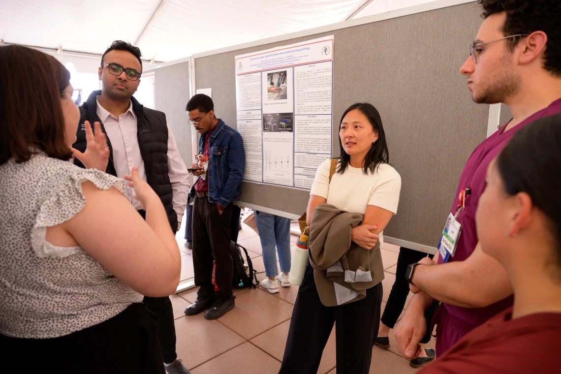 [Emergency Medicine’s Alli Min, MD, listens to a student talk about her research project at the COM-T 2025 Research Day poster session, while David Jordan, PhD (behind Dr. Min), checks his phone messages. A co-author on Jordan’s research poster, “Ultrasound shear wave speed of the transverse carpal ligament during pinching,” is the DOM Division of Rheumatology’s C. Kent Kwoh, MD, who also has been the U of A Arthritis Center director since 2013.]