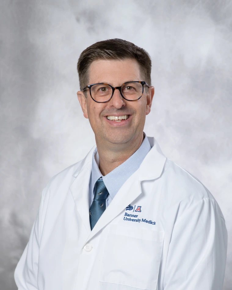 [A white man wearing glasses in physician's white coat with blue shirt and tie]
